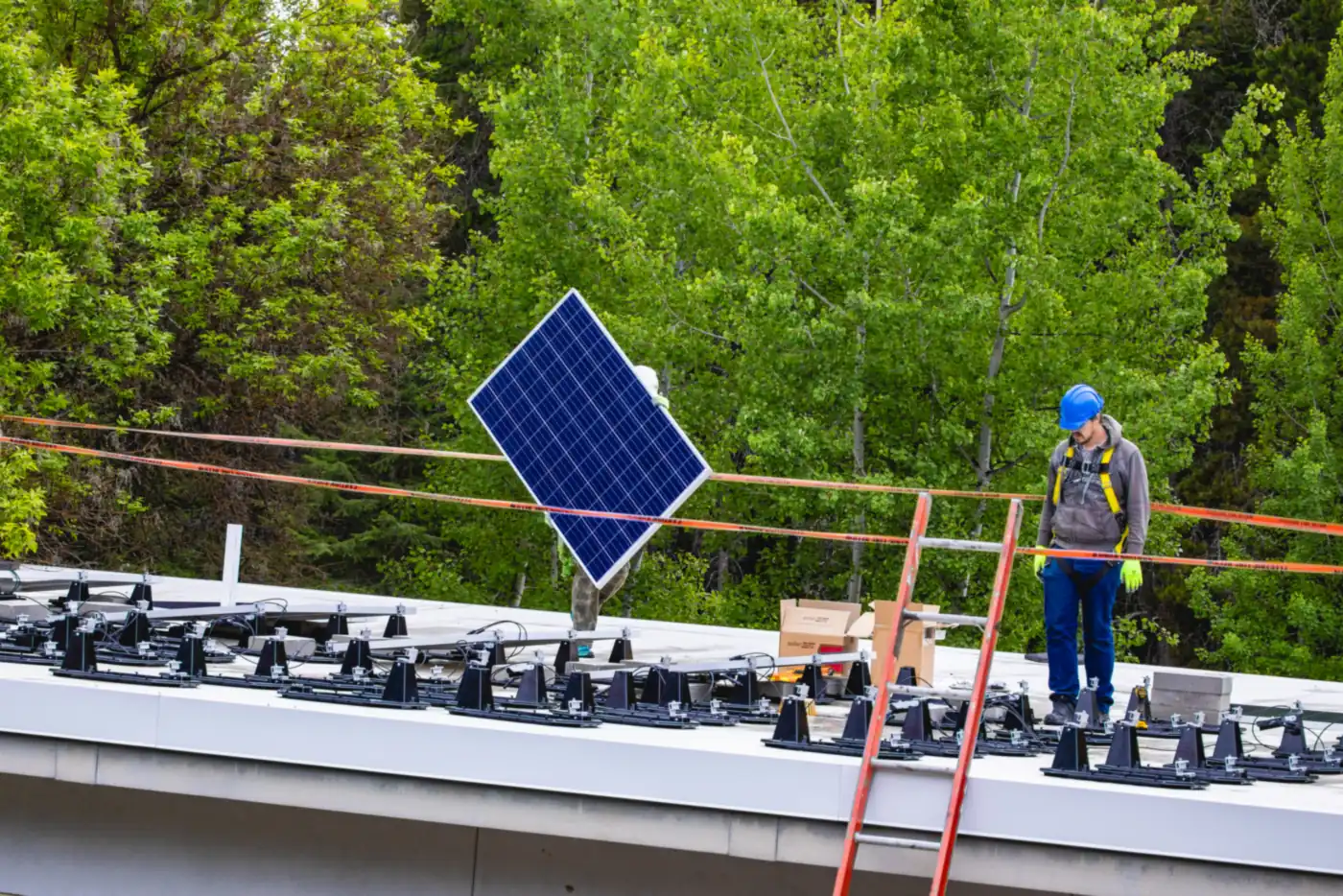Monteur mit Sicherungsgurt steht auf einem Flachdach zwischen Montagefüßen, ein Solarmodul wird hochgereicht (Glatten, Landkreis Freudenstadt)