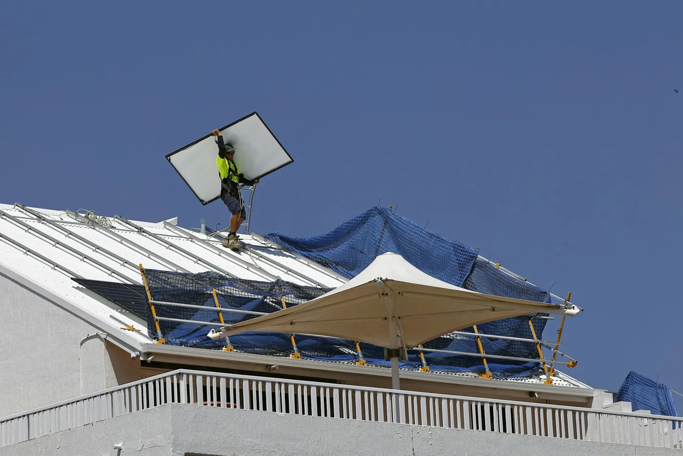 Monteur in Warnkleidung trägt ein Solarmodul über ein Blechdach mit Schutznetz und Geländer (Tschernitz, Wokrejs Sprjewja-Nysa)