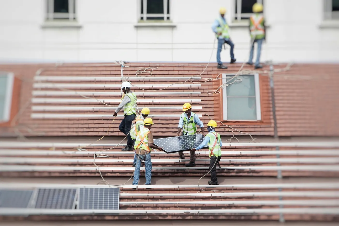 Montageteam mit Helmen und Sicherungsseilen trägt ein Solarmodul über die Schienen eines Ziegeldachs (Espenau, Landkreis Kassel)